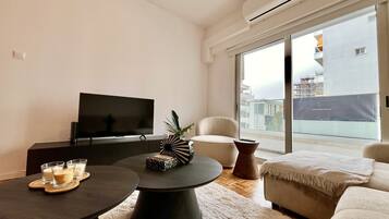 Ein modernes Wohnzimmer mit Flachbildfernseher, rundem Couchtisch, Sofa und großem Fenster mit Blick auf Gebäude.