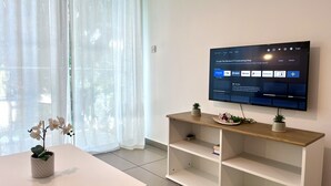 A modern living room with a flat-screen TV mounted on the wall, a white table with a potted plant, and a wooden shelf unit. - STAY Tropical Apartment (Protaras)
