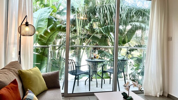 A room with a sofa, a table, and a balcony with chairs and plants. - STAY Tropical Apartment (Protaras)