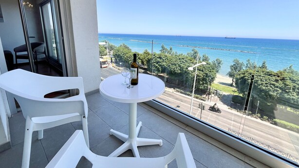 Un balcón con una mesa redonda, dos sillas blancas, una botella de vino y vistas al mar y la vegetación.