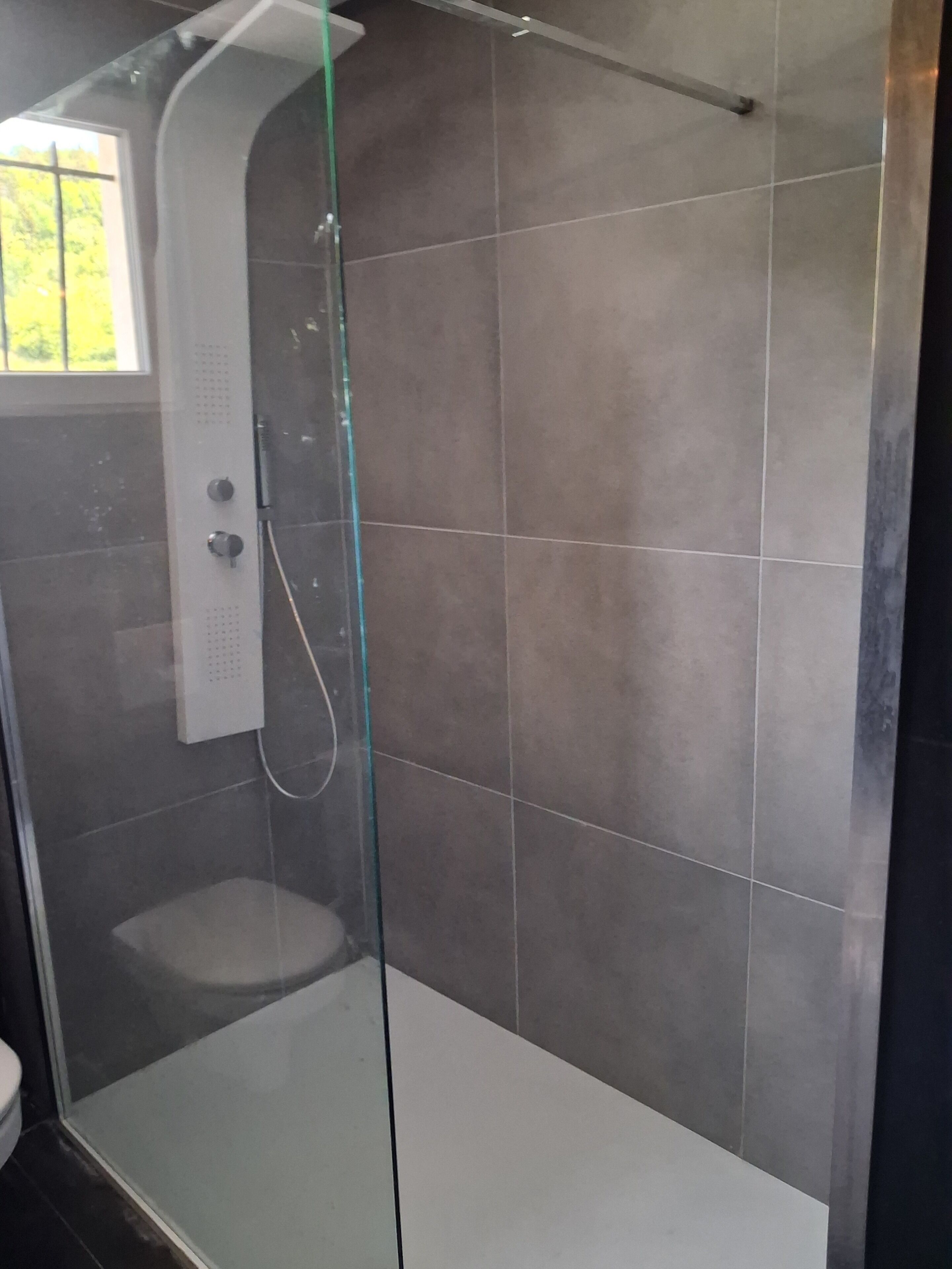 Eine begehbare Dusche mit Glaswänden, einer weißen Bedienfeld, gefliesten Wänden und einem sichtbaren Fenster.