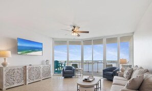 A spacious living room with a large window, a ceiling fan, and a flat-screen TV mounted on the wall. - Caribe Penthouse 6B by Liquid Life (Orange Beach)