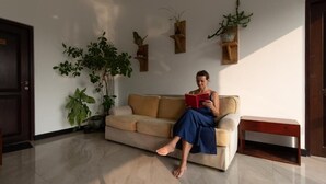 Una persona sentada en un sofá en una habitación con una puerta, una planta en maceta y estantes fijados a la pared.