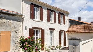 A two-story building with white walls and brown shutters, a stone wall on the left, and a tiled roof. - Country House “La Demeure D'Elles” with Private Terrace and Garden (Le Thou)