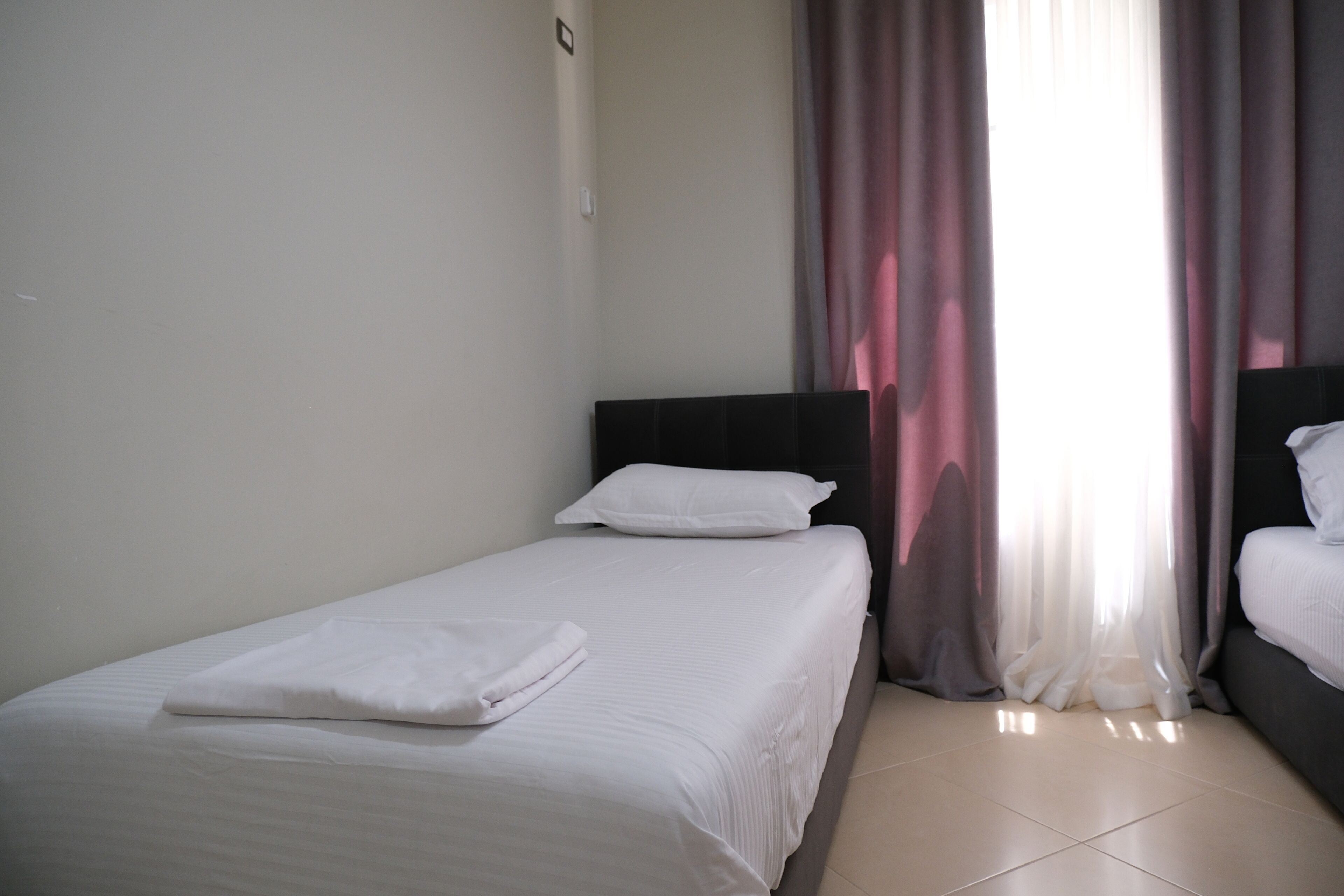 A hotel room with a single bed, a nightstand, a window with curtains, and a tiled floor.