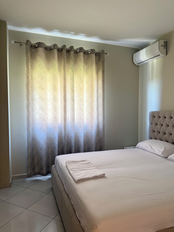 A bedroom with a bed, curtains, an air conditioning unit, and a tiled floor.