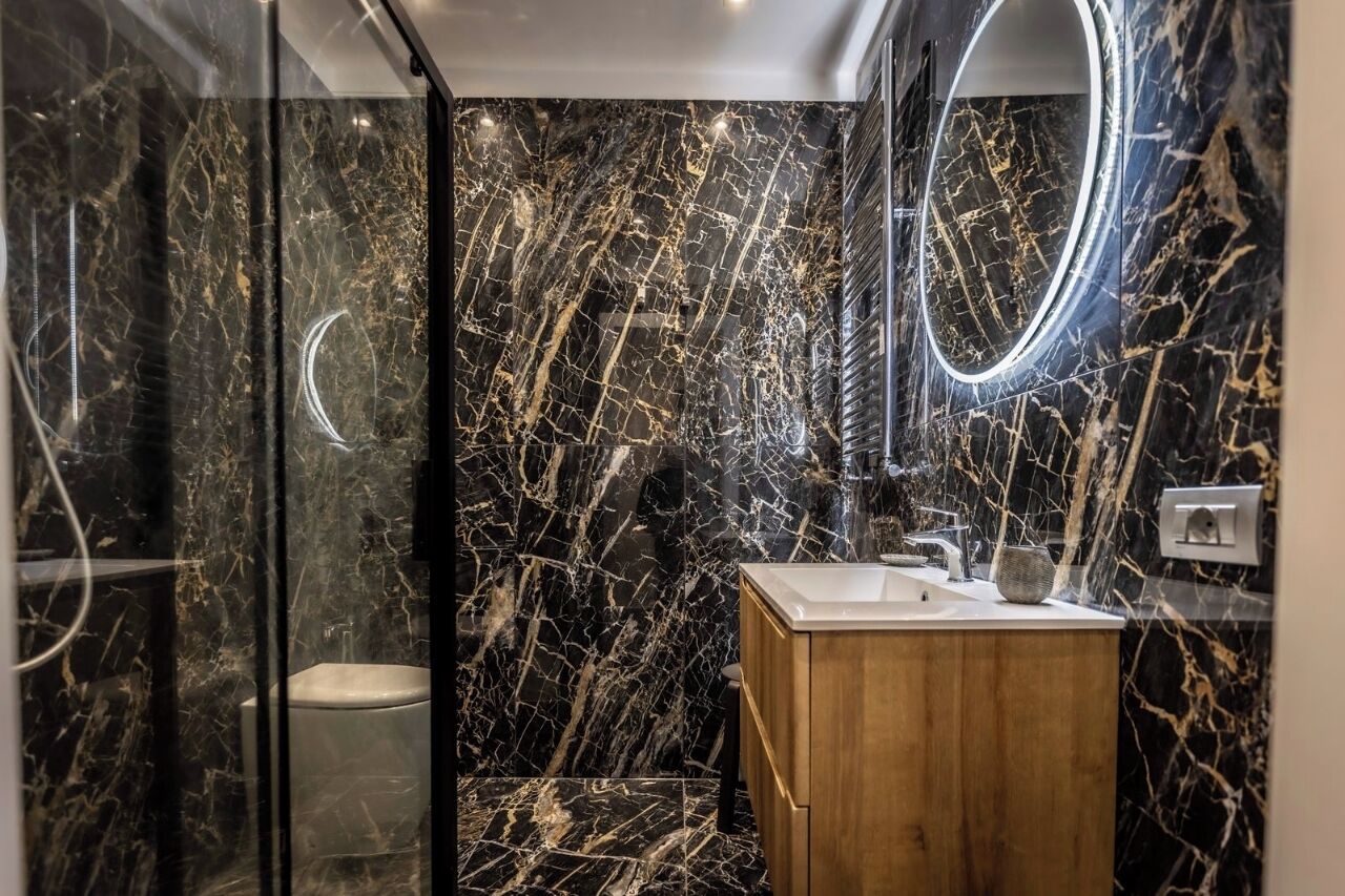 A bathroom with a marble wall, a wooden vanity, a toilet, and a mirror with a light.