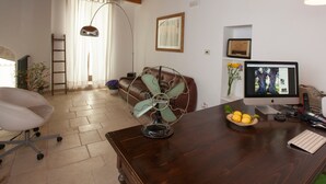 Ein Zimmer mit Holzboden, einem Schreibtisch mit Computer, einem Stuhl, einem Ventilator und einem Fenster mit Vorhängen.
