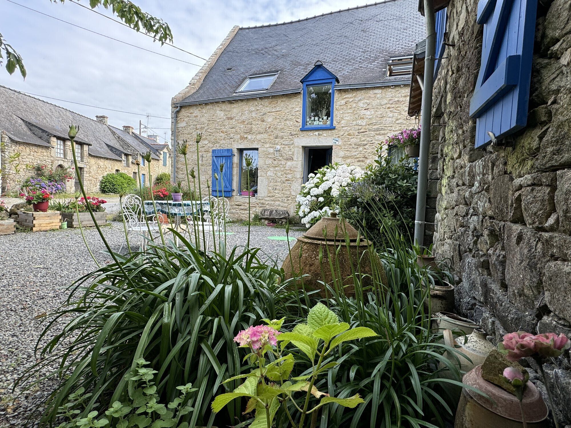 Un edificio de piedra con una ventana y contraventanas azules, rodeado por un patio de grava con plantas y flores en macetas.
