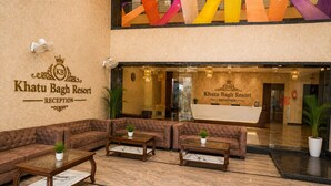 Un área de recepción con un letrero que indica Khatu Bagh Resort, un espacio de espera con sofás y mesas, y un ventanal grande.