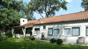 A white building with a red-tiled roof, a covered porch, and a well-maintained lawn. - Family Bungalow With Enclosed Garden (Koudekerke)
