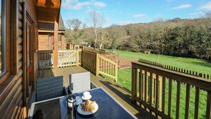 A wooden deck with outdoor furniture, a table with a coffee pot, cups, and croissants, and a view of a grassy area with trees and hills. - Trevano (Bodmin)