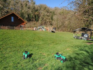 A grassy yard with two green toy tractors, a wooden playhouse, and a wooden structure with a roof. - Trevano (Bodmin)