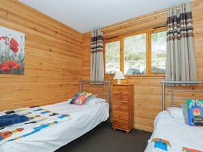A wooden cabin room with two beds, a dresser, a window with curtains, and a painting on the wall. - Trevano (Bodmin)