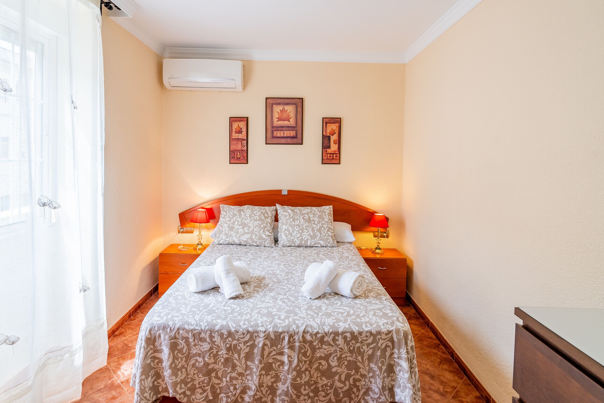 A hotel room with a bed, two bedside lamps, a nightstand, a dresser, and a window with curtains.