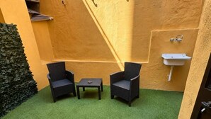 A small outdoor area with artificial grass, two wicker chairs, a small table, and a white sink against a yellow wall.