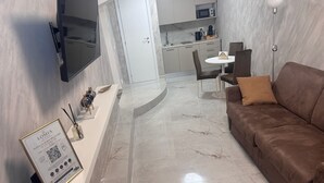 A modern living room with a flat-screen TV, a brown sofa, a white coffee table, and a kitchen area in the background.