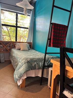 A room with a bed, a desk, a window with curtains, and a view of greenery outside.