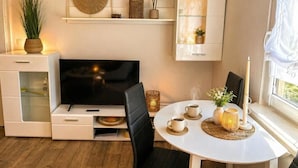 Un salon moderne avec un téléviseur à écran plat, une table à manger avec des chaises, un meuble blanc avec des portes en verre et une fenêtre avec des rideaux.