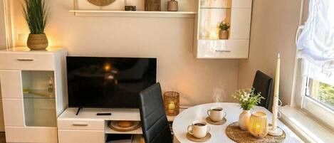 A modern living room with a flat-screen TV, a dining table with chairs, a white cabinet with glass doors, and a window with curtains.