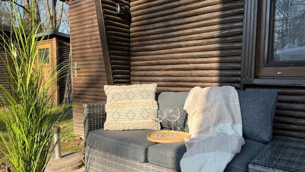 Un canapé en osier, agrémenté de coussins et d’un plateau, est placé à l’extérieur d’une cabane en bois dotée d’une fenêtre.