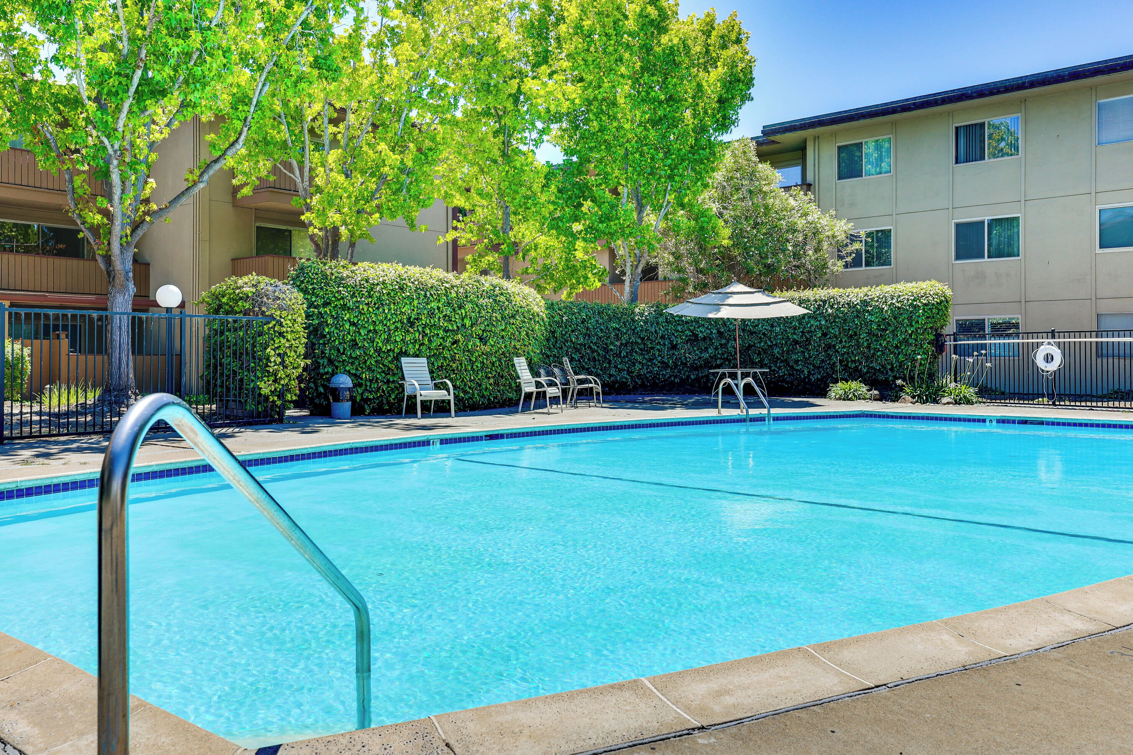 A clear swimming pool with a metal ladder, surrounded by a fence, and adjacent to a building with multiple windows.