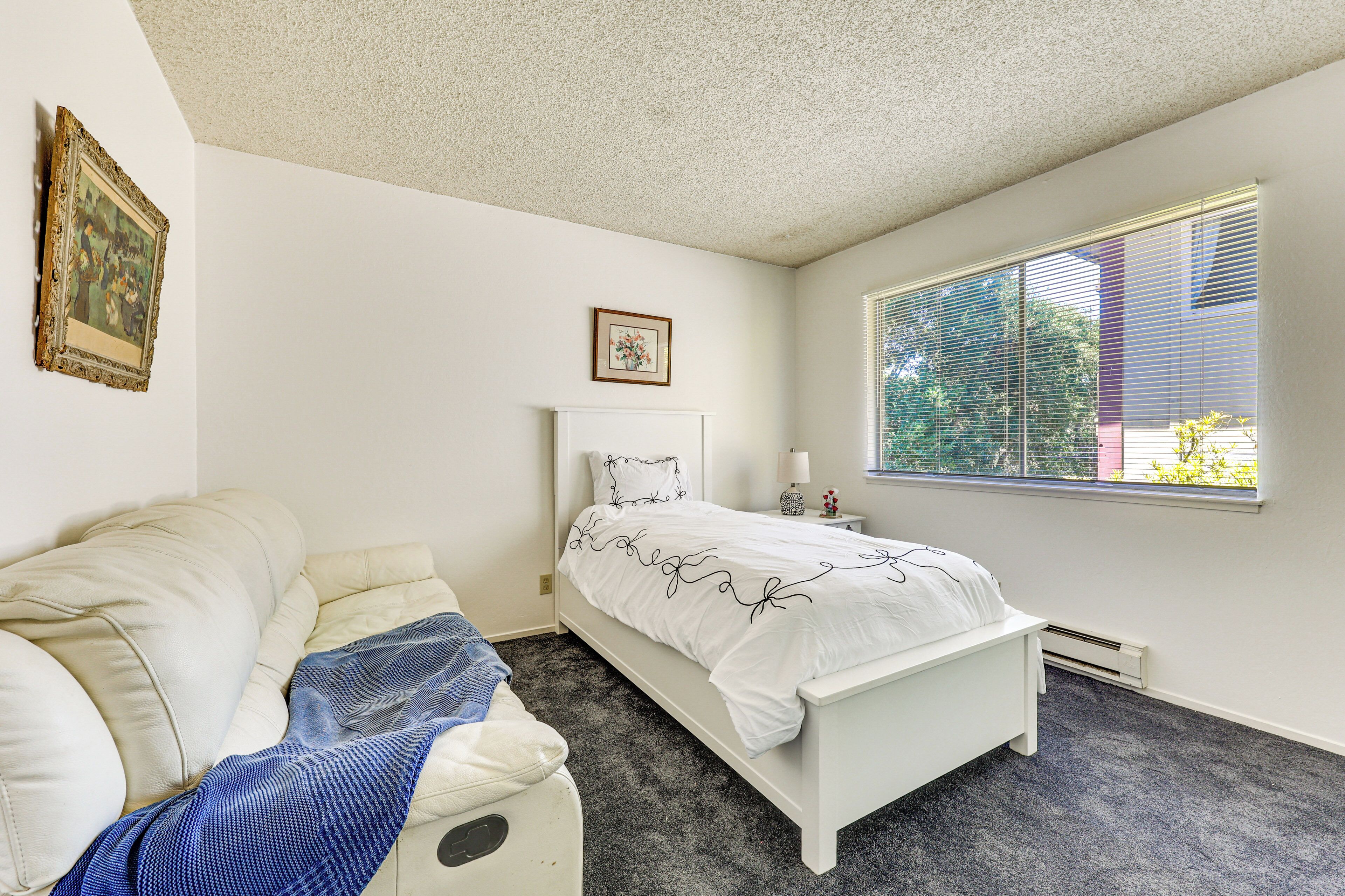 A bedroom with a white bed, a white bench, a window with blinds, and a picture on the wall.