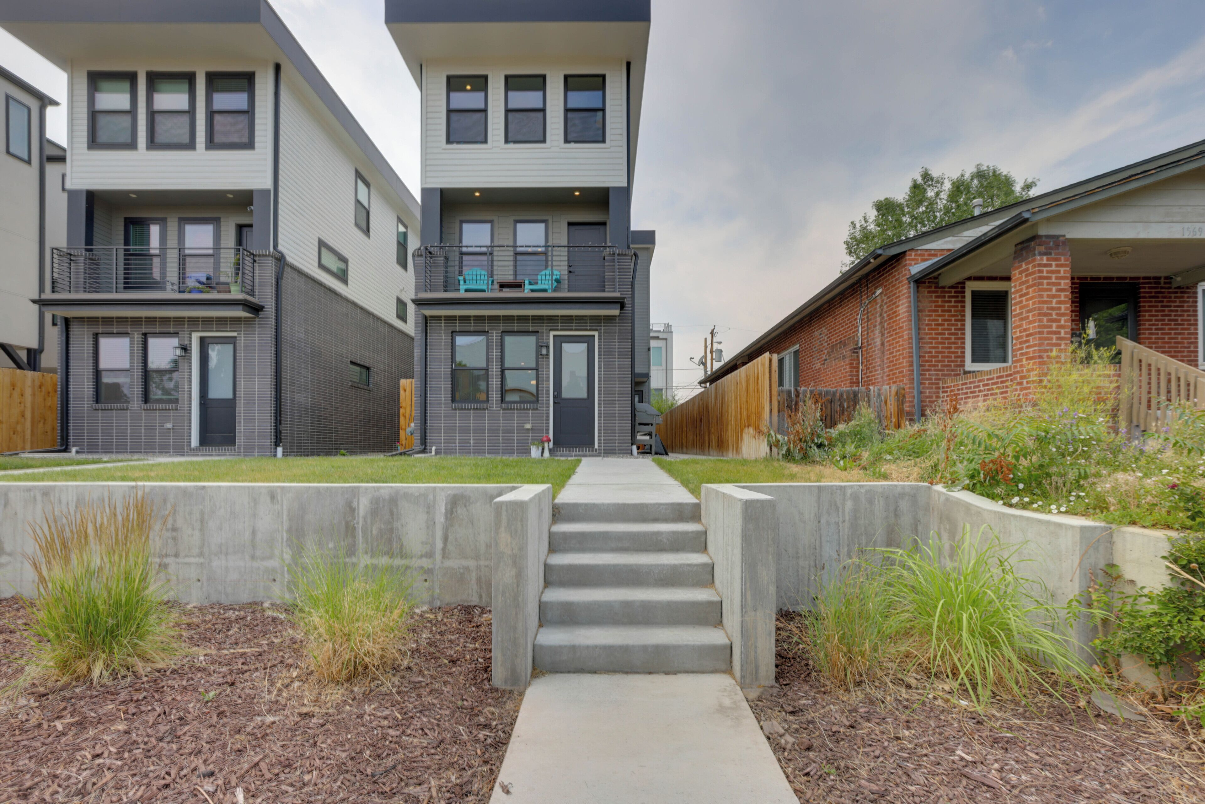 A modern multi-story residential building with a central staircase leading to the entrance, surrounded by landscaped areas with grass and shrubs.