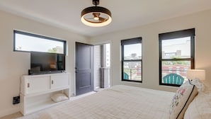 Une chambre à coucher avec un lit, un téléviseur posé sur une commode blanche, un ventilateur de plafond et de grandes fenêtres.