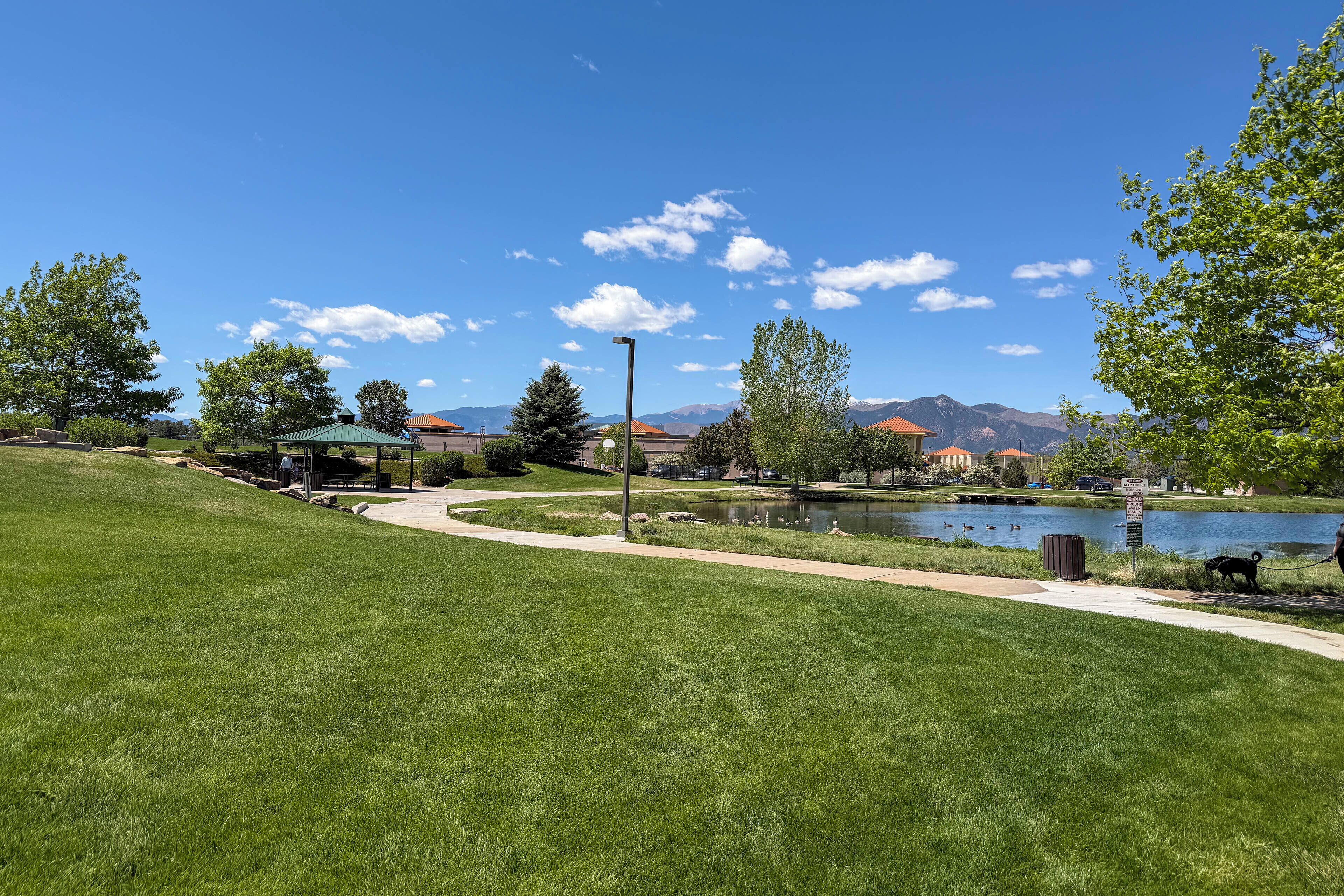 A park with a walking path, a gazebo, a pond, and mountains in the background.