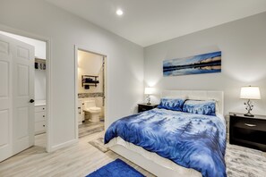 A bedroom with a bed, a nightstand, a wall-mounted lamp, a bathroom visible through an open door, and a painting on the wall. - Explore The Springs! Family-friendly Basement Unit (Colorado Springs)