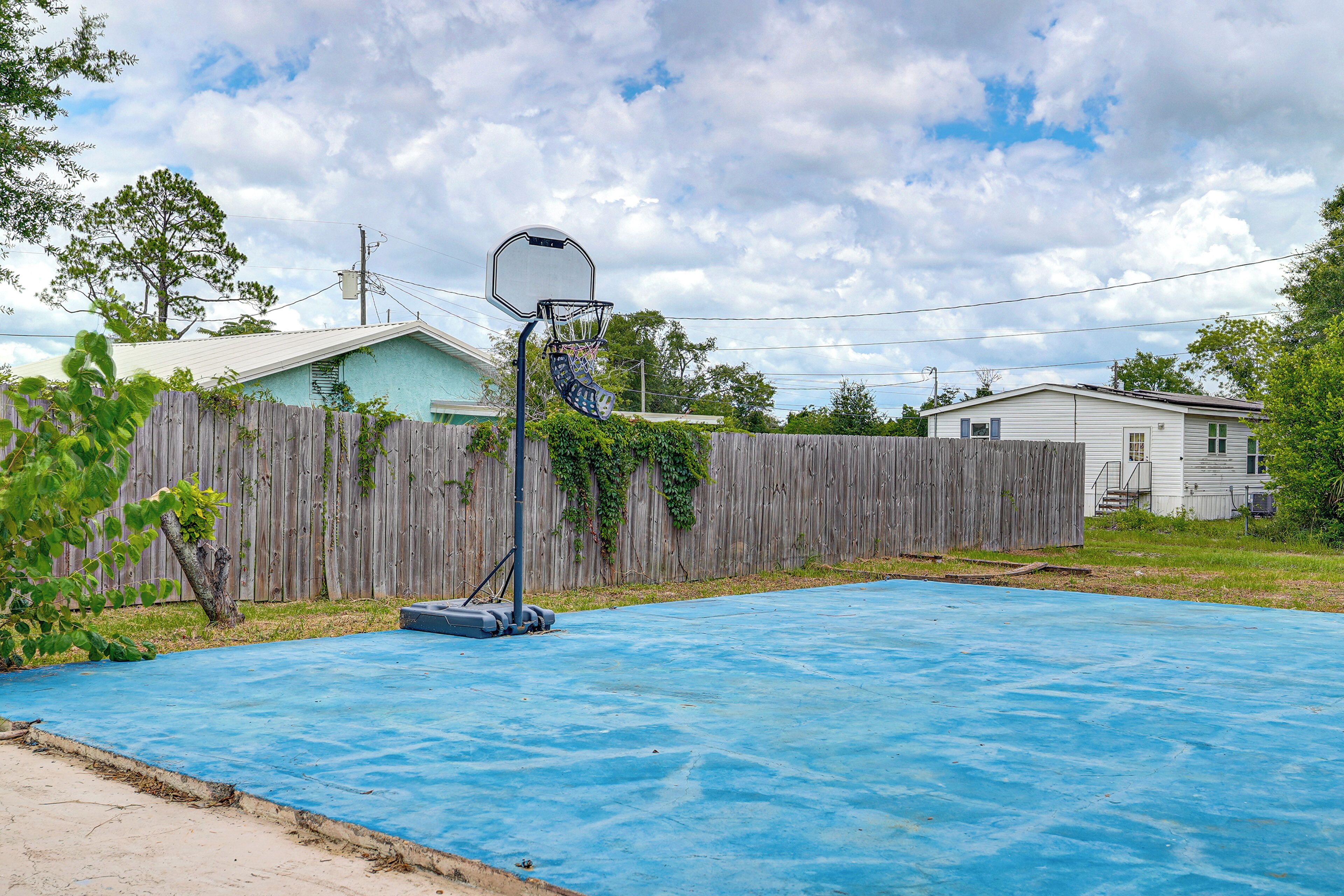 Un campo da basket blu all'aperto con canestro, circondato da una recinzione di legno e case sullo sfondo.
