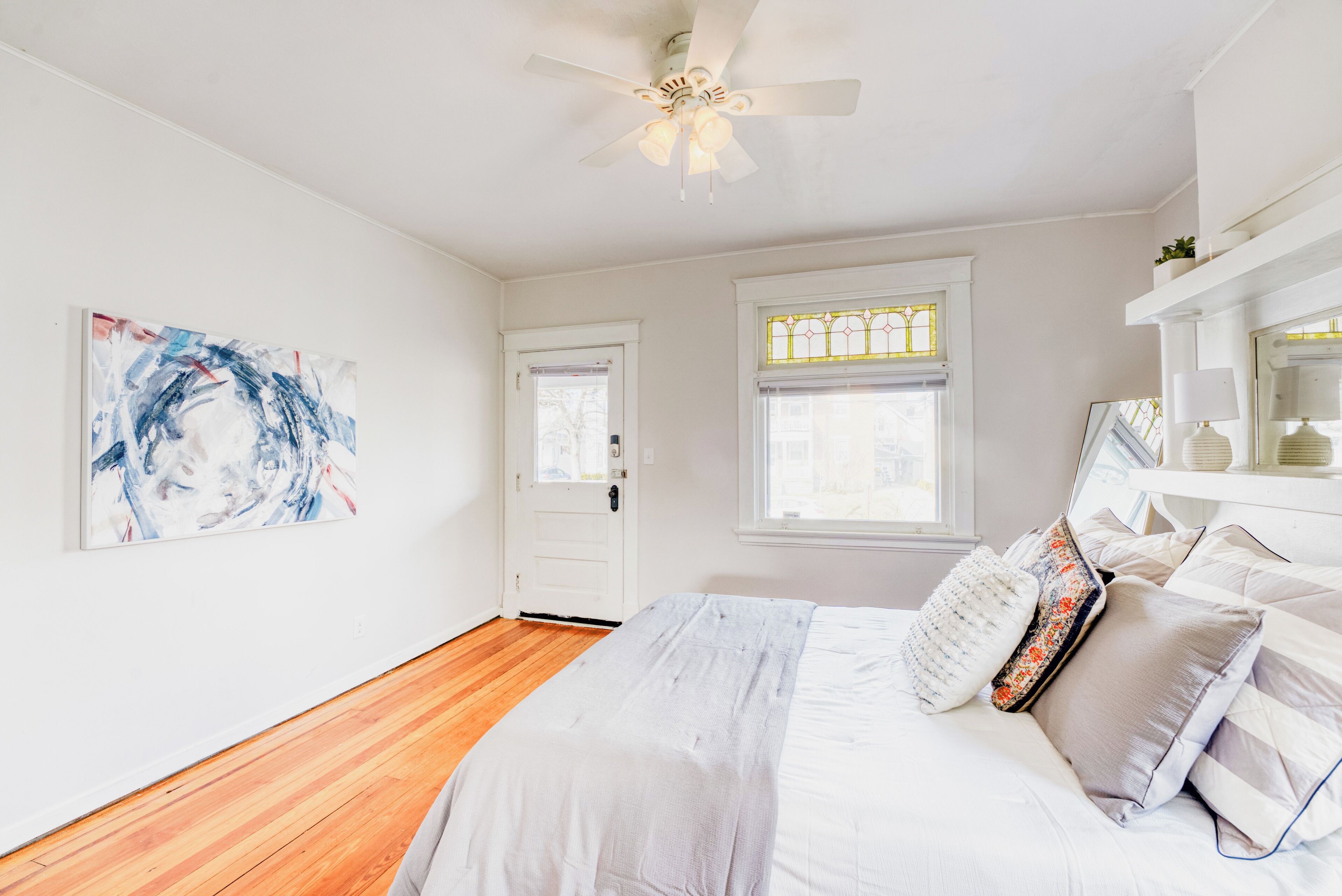 Une chambre avec un plancher en bois, un lit avec une literie blanche, un ventilateur de plafond, une porte et une fenêtre ornée de vitraux.