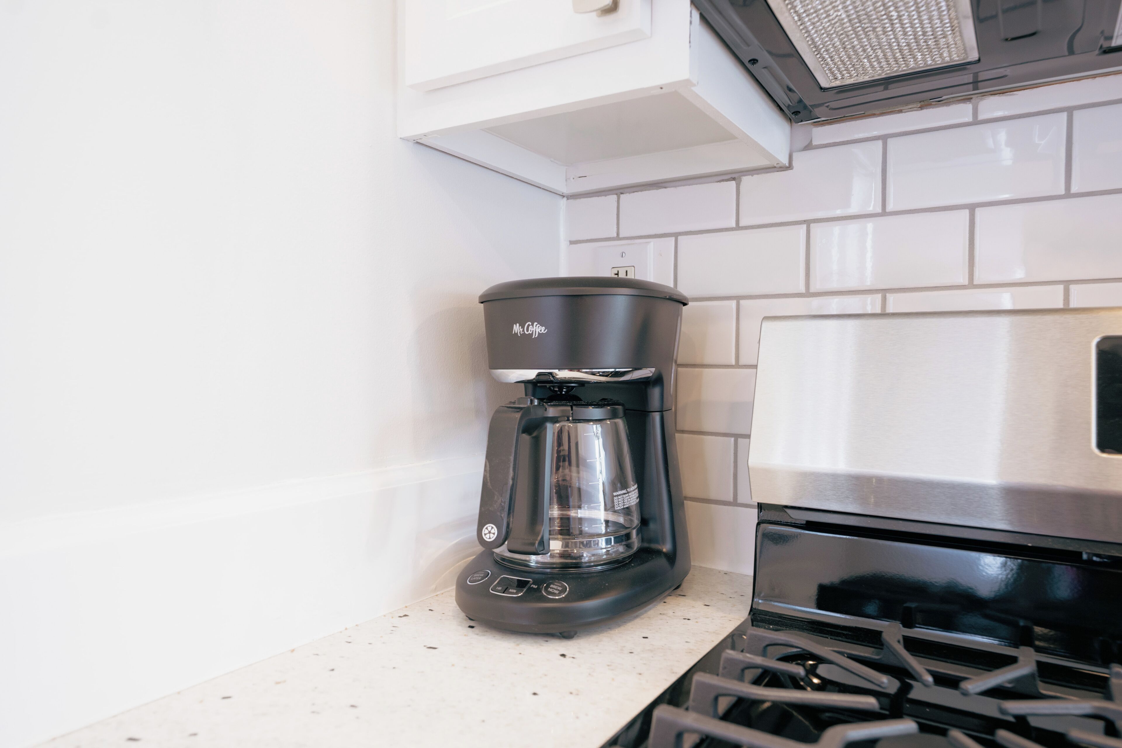 Une cuisine équipée d’une machine à café, d’une cuisinière et de murs recouverts de carreaux blancs.