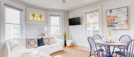 A living room with a white sofa, a dining table with chairs, a ceiling fan, and a TV mounted on the wall.