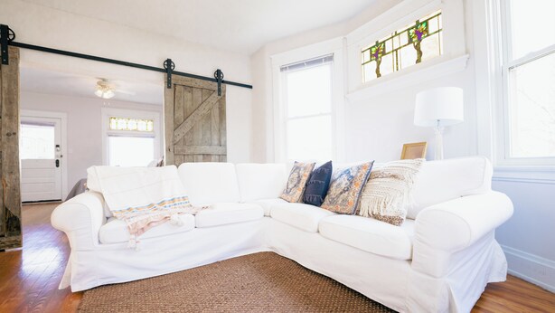 A white sectional sofa with a brown rug, a wooden barn door, and a large window with stained glass. - Walkable Oakley Neighborhood Home in Cincinnati! (Cincinnati)