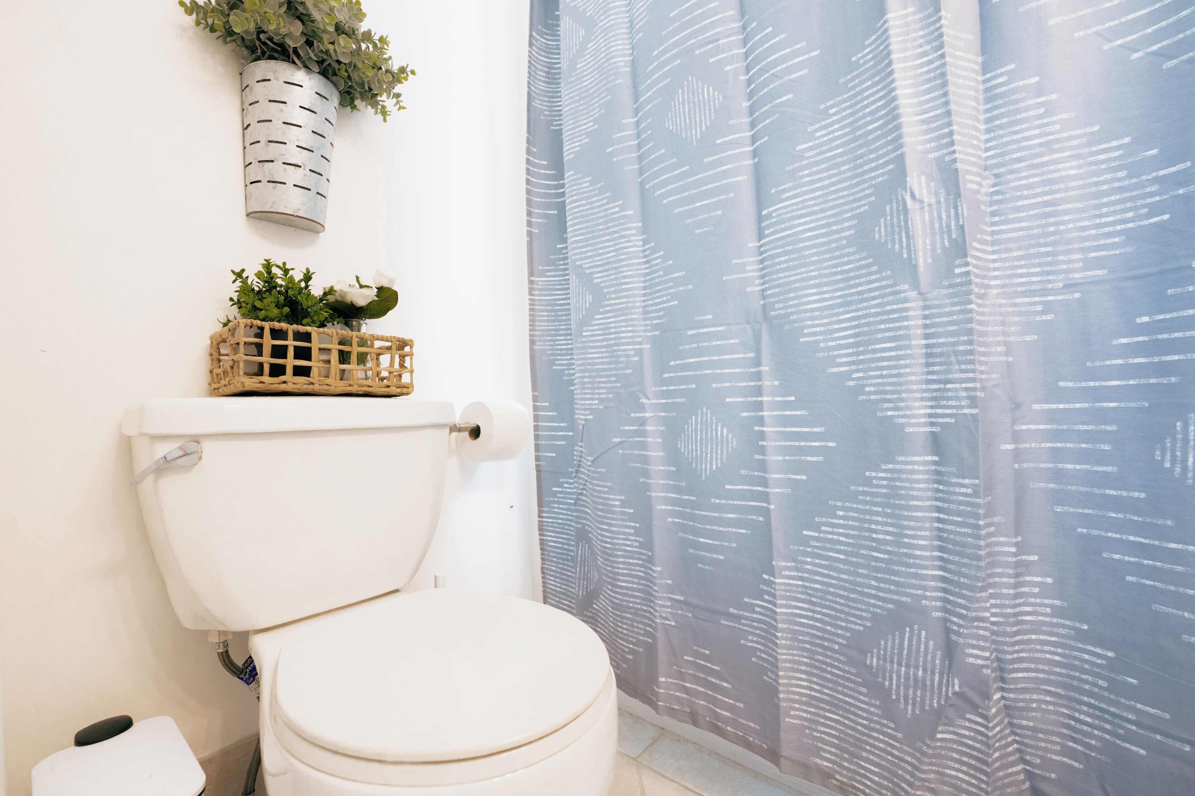 A toilet with a white tank, a wall-mounted planter with greenery, and a patterned shower curtain.