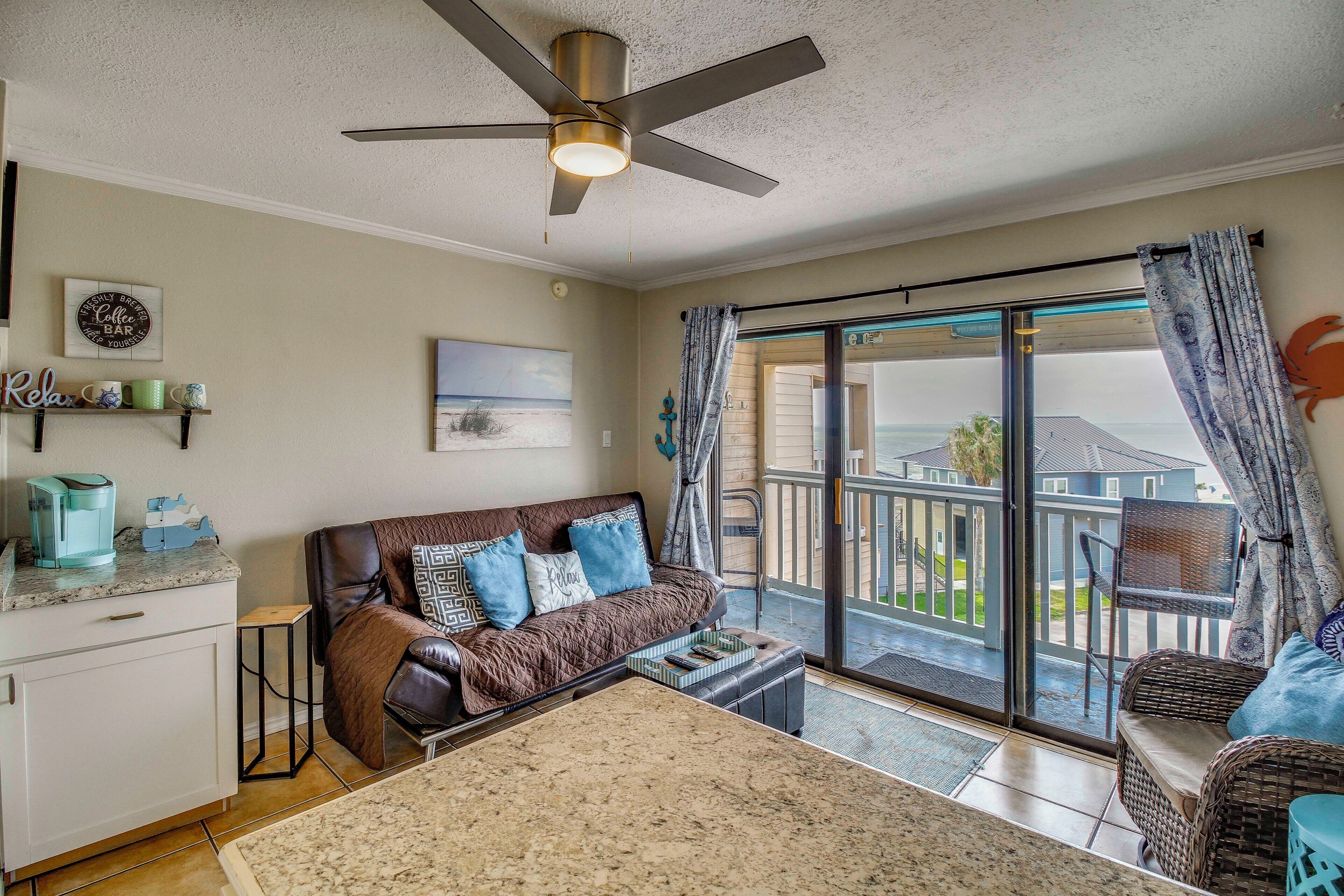 A living room with a sofa, a coffee table, a ceiling fan, a sliding glass door to the outside, and a kitchen area with a countertop and cabinets.