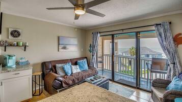 A living room with a sofa, a coffee table, a ceiling fan, a sliding glass door to the outside, and a kitchen area with a countertop and cabinets.