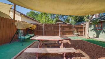 A backyard with a wooden table, a swing set, and a shaded area.