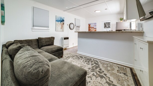 Ein Wohnzimmer mit Sofa, einem gemusterten Teppich, einem Flachbildfernseher, einer kleinen Küchenzeile und einer Wanduhr.
