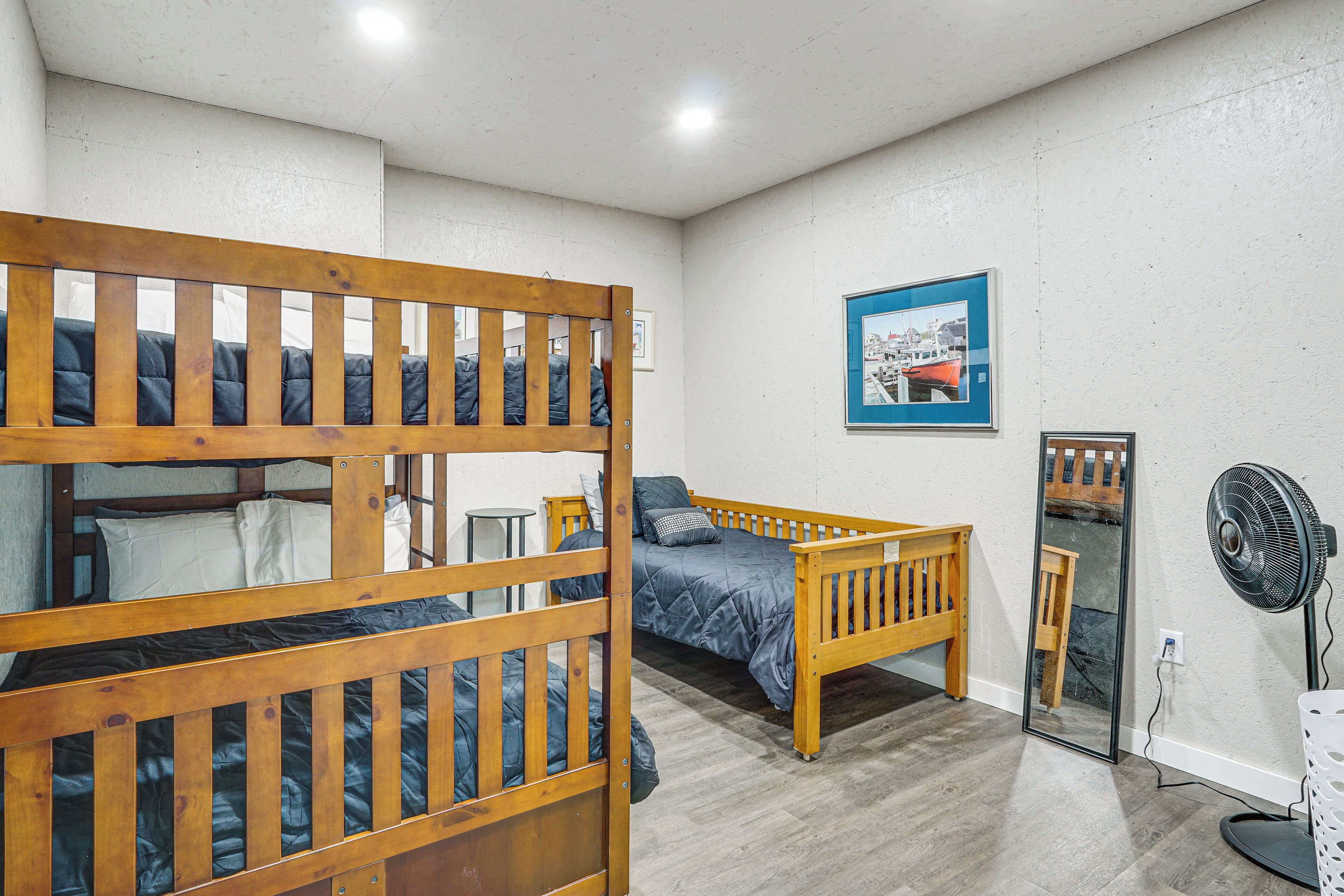A room with bunk beds, a single bed, a small table, a chair, a mirror, a fan, and a framed picture on the wall.