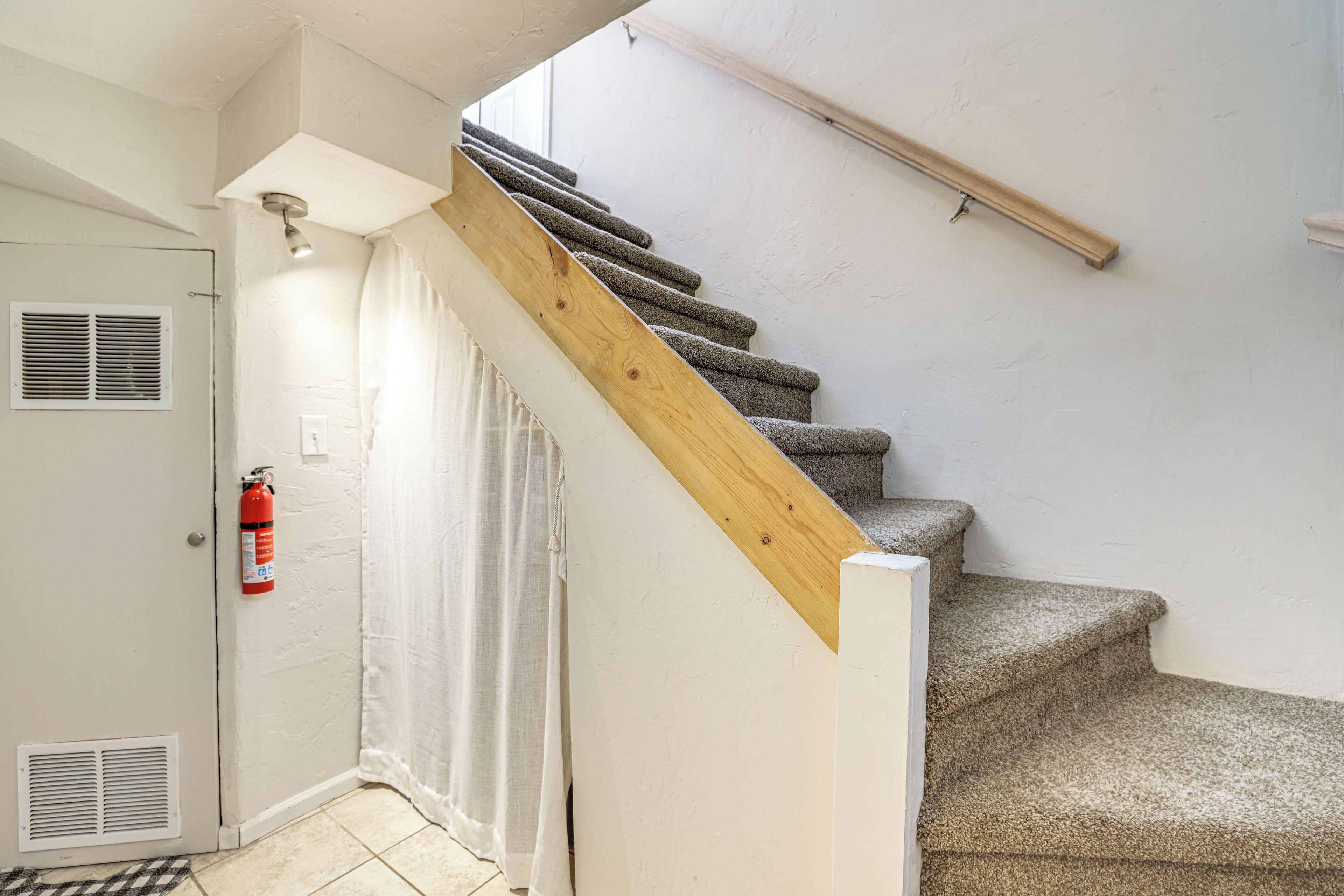 Una escalera con peldaños alfombrados, barandilla de madera, extintor, puerta con rejilla de ventilación y cortina.