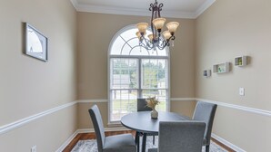 Un coin repas avec une table ronde, quatre chaises, un lustre, une fenêtre avec des stores et un tableau encadré au mur.