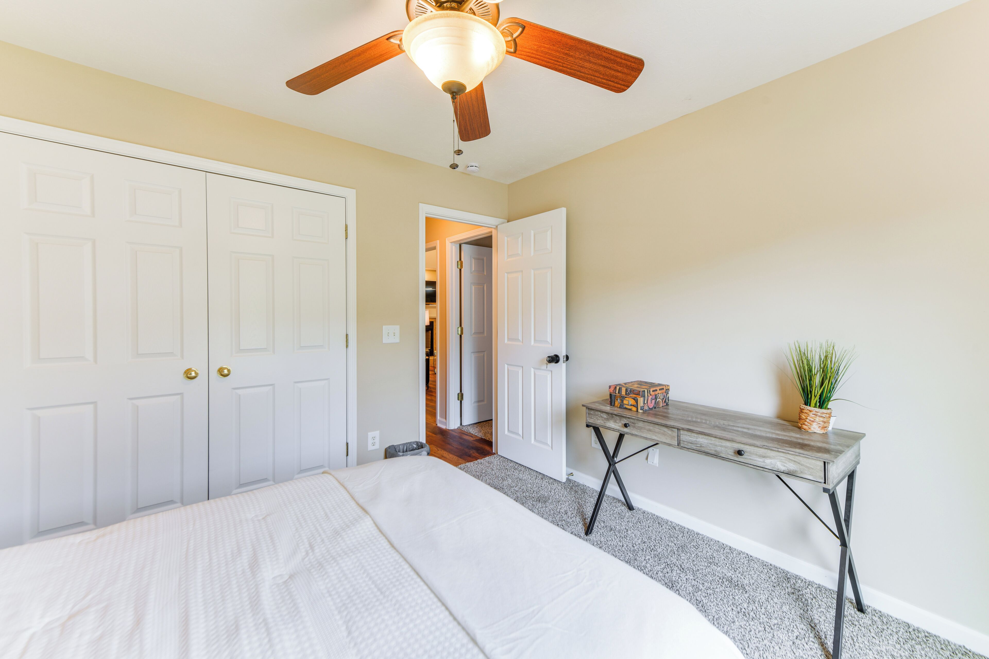 A bedroom with a bed, a ceiling fan, a wooden desk, and a door leading to another room.