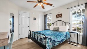 A bedroom with a bed, a ceiling fan, a window with curtains, a bedside table, and a chair. - Near Eats, Shops, Golf & Beach: Oak Island Suite! (Oak Island)