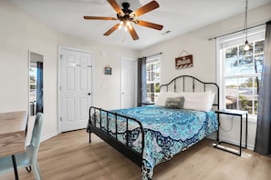 Quarto com cama, ventoinha no teto, janela com cortinas, mesa de cabeceira e uma cadeira.