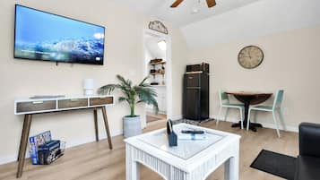 A modern living room with a flat-screen TV, a dining area with a table and chairs, a kitchen with a refrigerator, and a ceiling fan.