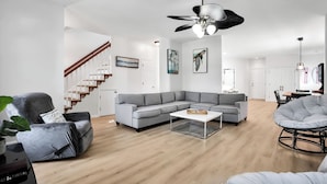Ein modernes Wohnzimmer mit einem grauen Ecksofa, einem runden Sessel, einem Couchtisch, einer Treppe und einem Deckenventilator.