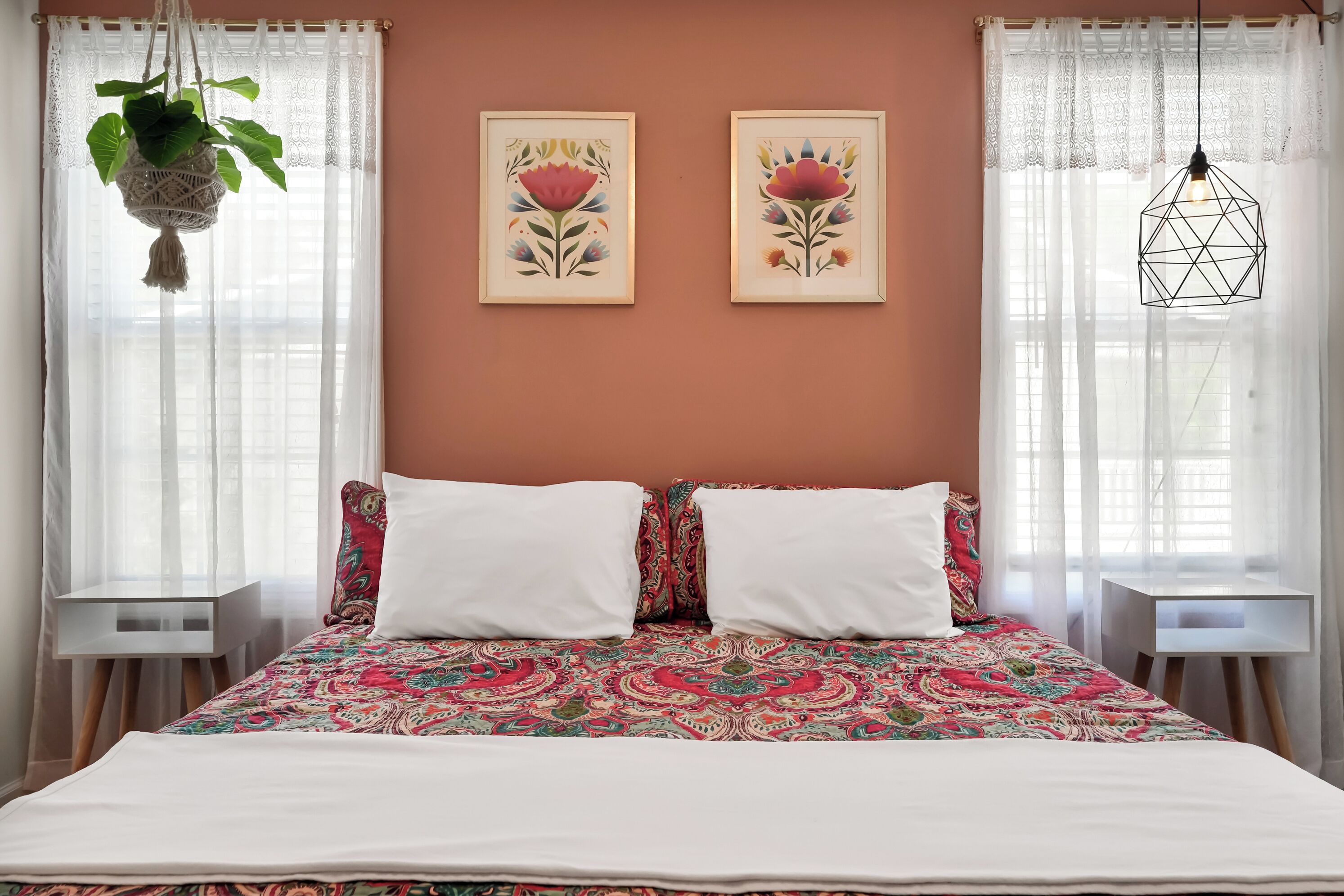 Ein Schlafzimmer mit einem Bett, zwei Kissen, einer hängenden Pflanze und zwei gerahmten floralen Kunstwerken an der Wand.